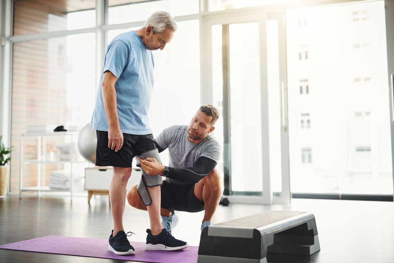 Senior, man and physiotherapist with knee bandage for support, circulation or compression for healing. Fitness, physiotherapy and elderly person with joint wrap, mobility training and exercise in gym