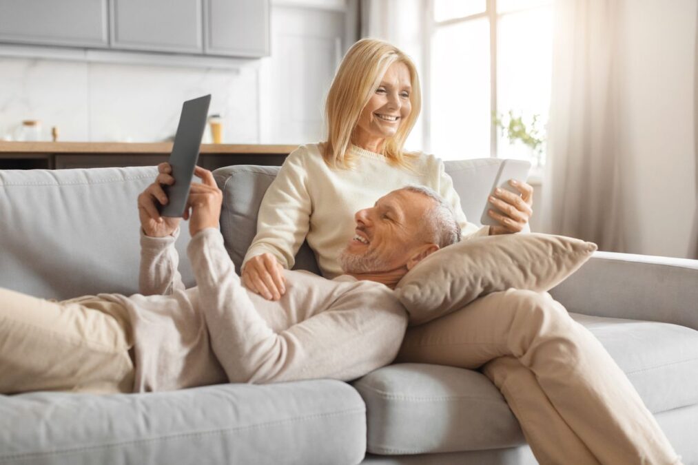 Senior man laughs while browsing a tablet as his partner enjoys her drink, both sharing a fun moment