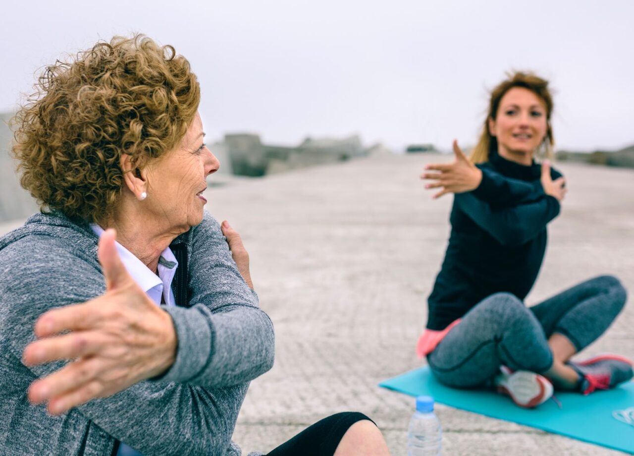 Senior woman stretching with female coach by sea pier